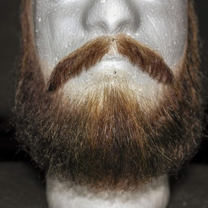 Puede incluir: Primer plano de una barba y bigote falsos. La barba es marrón y el bigote es marrón claro. La barba está peinada en una barba corta y completa con bigote.