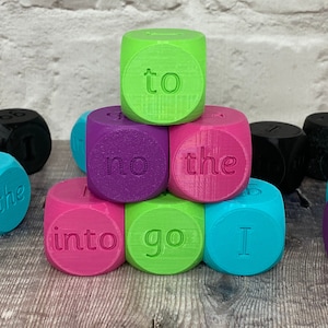 May include: A set of colourful dice with words such as "go", "the", "to", "no", "into", and "I" printed on them. The dice are arranged in a pyramid shape on a wooden surface.