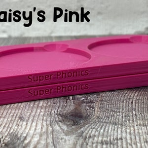 Pu&ograve; includere: Due vassoi di plastica rosa con incavi rotondi, impilati uno sopra l'altro. I vassoi sono etichettati "Super Phonics" in testo nero.