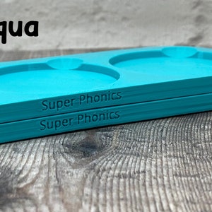 Pu&ograve; includere: Due vassoi in plastica blu turchese con tre incavi circolari. I vassoi sono impilati uno sopra l'altro. Il testo "Super Phonics" &egrave; stampato sul fondo di ogni vassoio.