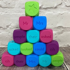May include: A pyramid of multi-coloured dice featuring words and a star. The dice are in shades of pink, purple, blue, green, and teal. The words include "Mr", "Mrs", "people", "their", "asked", "looked", "could", and "oh".