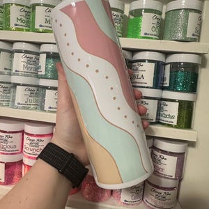May include: A white tumbler with a pastel wave design in pink, mint green, and beige, accented with small gold dots. The tumbler is cylindrical and appears to be held in a person's hand. Jars of glitter are visible in the background.