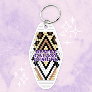 Puede incluir: Llavero de motel blanco con un patrón geométrico negro, blanco y marrón. El texto "DESERT GALAXY DESIGNS" está impreso en morado en el llavero.