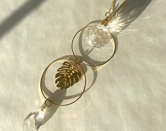Puede incluir: Un atrapasoles decorativo con una cadena dorada y detalles circulares. Cuenta con una bola de cristal transparente, una hoja de monstera dorada y un colgante de cristal en forma de lágrima. El diseño está suspendido de una cadena dorada.