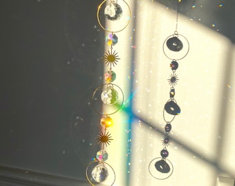 Puede incluir: Un atrapasol de cristal con anillos dorados y cristales en forma de estrella cuelga de un techo, proyectando luz arcoíris en una pared blanca.