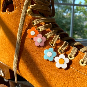 May include: Close-up of a tan roller skate with three flower charms in orange, pink, light blue, and white. The skate has tan laces and a tan suede exterior. The charms are attached to the laces.