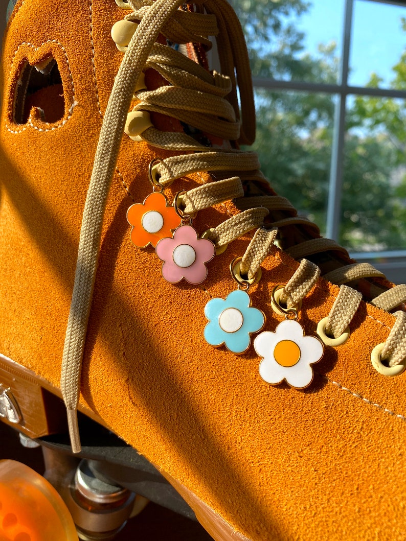 May include: Close-up of a tan roller skate with three flower charms in orange, pink, light blue, and white. The skate has tan laces and a tan suede exterior. The charms are attached to the laces.