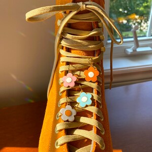 May include: Close-up of a mustard yellow roller skate with matching laces. Four flower-shaped earrings in pink, orange, blue, and white hang from the laces. The skate is made of suede.