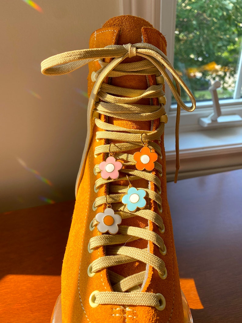 May include: Close-up of a mustard yellow roller skate with matching laces. Four flower-shaped earrings in pink, orange, blue, and white hang from the laces. The skate is made of suede.