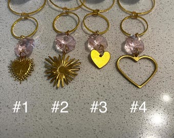 Op de afbeelding: Vier goudkleurige wijnbedels met verschillende hangerontwerpen. Elke bedel heeft een gouden ring, een lichtroze kristal en een gouden hanger. De hangers omvatten een hart, een zonnestraal en een hartvormige omtrek.