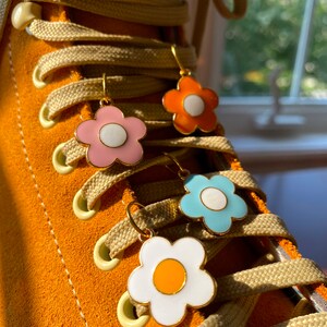 May include: Close-up of four flower-shaped earrings in pink, orange, blue, and white with gold-colored hoops. The earrings are hanging on the laces of an orange suede boot. The laces are beige.
