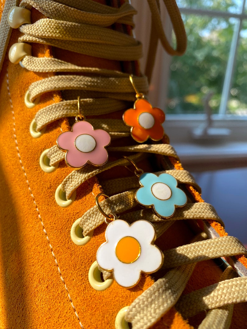 May include: Close-up of four flower-shaped earrings in pink, orange, blue, and white with gold-colored hoops. The earrings are hanging on the laces of an orange suede boot. The laces are beige.