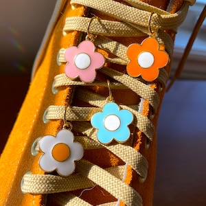 May include: Close-up of four flower-shaped earrings in pink, orange, blue, and white with gold-colored hooks. The earrings are hanging on the laces of a bright orange sneaker. The earrings have a white center and a gold-colored outline.
