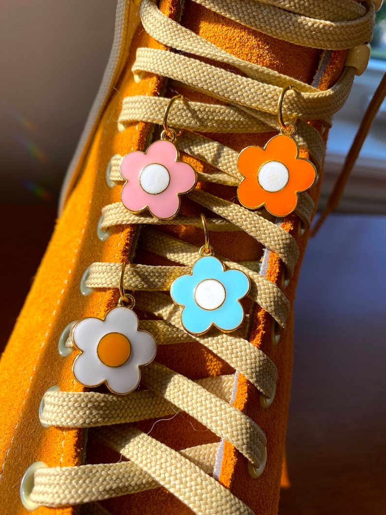 May include: Close-up of four flower-shaped earrings in pink, orange, blue, and white with gold-colored hooks. The earrings are hanging on the laces of a bright orange sneaker. The earrings have a white center and a gold-colored outline.