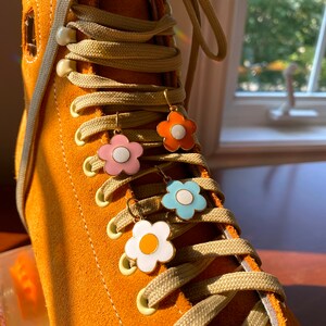 May include: Close-up of an orange suede roller skate with beige laces and four flower-shaped earrings in pink, orange, blue, and white. The earrings have gold-colored hooks and white centers. The skate is in focus, with a blurred background.
