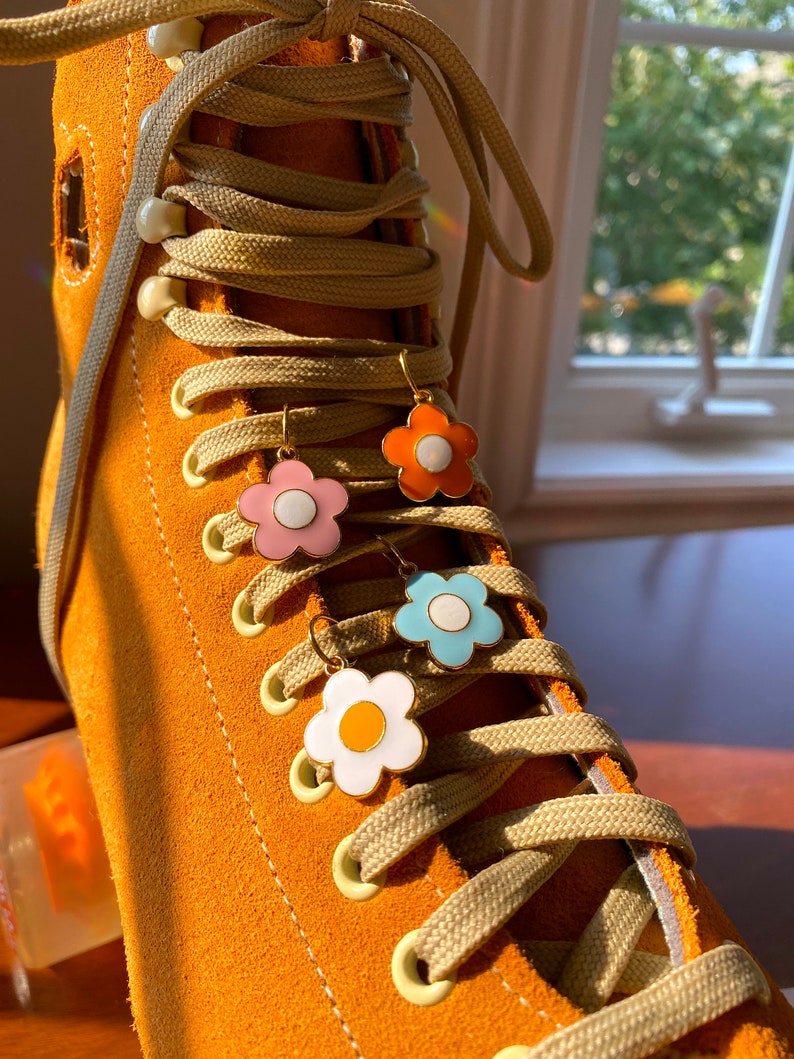 May include: Close-up of an orange suede roller skate with beige laces and four flower-shaped earrings in pink, orange, blue, and white. The earrings have gold-colored hooks and white centers. The skate is in focus, with a blurred background.