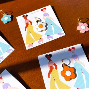 May include: Flower-shaped enamel charms in blue, white, pink, and orange with gold-colored hoops. The charms are displayed on cards with illustrations of figures roller skating. The text "jvmakesthings" is visible on the cards.