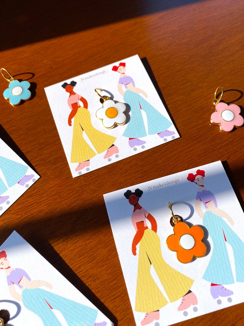 May include: Flower-shaped enamel charms in blue, white, pink, and orange with gold-colored hoops. The charms are displayed on cards with illustrations of figures roller skating. The text "jvmakesthings" is visible on the cards.