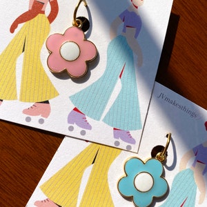 May include: Two flower-shaped earrings with gold-colored trim. One earring is light pink with a white center, and the other is light blue with a white center. The earrings are displayed on cards with illustrations of figures in retro roller skates. The text "Jvmakesthings" is visible.