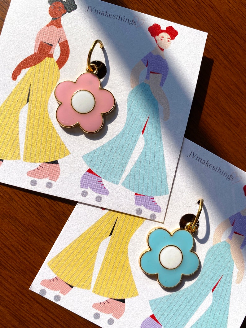 May include: Two flower-shaped earrings with gold-colored trim. One earring is light pink with a white center, and the other is light blue with a white center. The earrings are displayed on cards with illustrations of figures in retro roller skates. The text "Jvmakesthings" is visible.