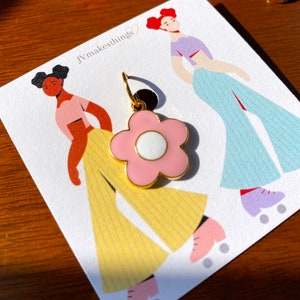 May include: A pink flower-shaped earring with a white center and gold-colored accents. The earring is displayed on a card with illustrations of figures roller skating. The card has the text "JVmakesthings".