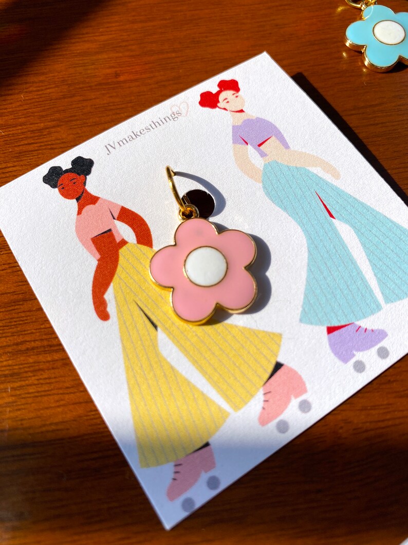 May include: A pink flower-shaped earring with a white center and gold-colored accents. The earring is displayed on a card with illustrations of figures roller skating. The card has the text "JVmakesthings".