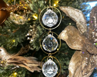 Puede incluir: Adorno navideño con tres esferas de cristal transparente suspendidas en aros dorados. Las esferas están facetadas, reflejando la luz. El adorno cuelga de una cadena dorada, sobre un fondo de árbol de Navidad decorado con follaje dorado y verde.