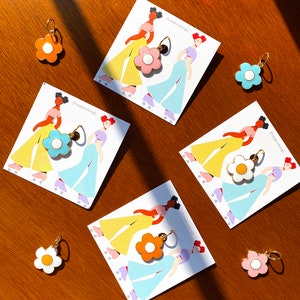May include: A collection of flower-shaped earrings in various colors, including orange, pink, blue, and white, with gold-colored hoops. The earrings are displayed on cards featuring illustrations of women roller skating. The brand name "jvmakesthings" is printed on the cards.