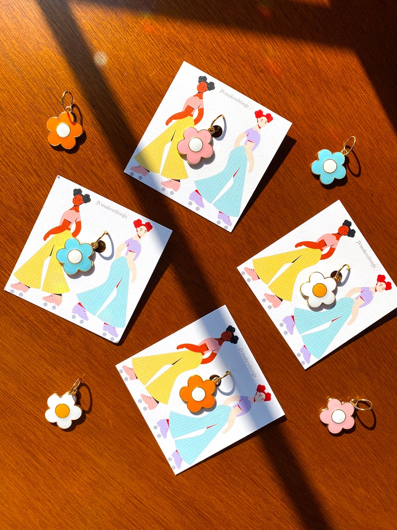 May include: A collection of flower-shaped earrings in various colors, including orange, pink, blue, and white, with gold-colored hoops. The earrings are displayed on cards featuring illustrations of women roller skating. The brand name "jvmakesthings" is printed on the cards.