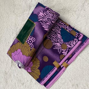 May include: Purple and pink fabric with a floral pattern and gold accents. The fabric is folded in half and has a label that says "Veritable Wax Hollandais Guaranteed Dutch".