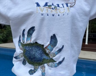 Może przedstawiać: Biała bluza z wyhaftowanym wzorem niebiesko-zielonego kraba na dole z przodu. Na klatce piersiowej znajduje się napis MAUI HAWAII wyhaftowany niebieską i beżową nicią, a nad nim mała grafika przedstawiająca palmy i żaglówkę.
