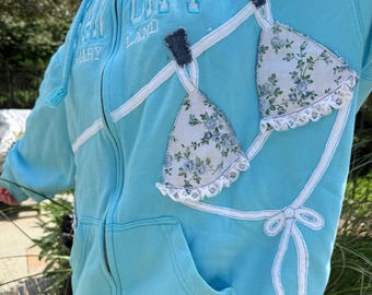 Może przedstawiać: Jasnoniebieska bluza z kapturem zapinana na zamek, z wyhaftowaną grafiką bikini na sznurku do bielizny. Góra bikini wykonana jest z białej tkaniny w niebieski kwiatowy wzór z białym koronkowym wykończeniem. Napis OCEAN CITY MARYLAND jest wyhaftowany na niebiesko.