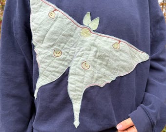 Może przedstawiać: Granatowa bluza z dużym, pikowanym naszywką w kształcie ćmy w kolorze jasnej zieleni z przodu. Ćma posiada haftowane detale na skrzydłach, w tym małe okrągłe oznaczenia w kolorze żółtym i brązowym oraz teksturowaną, przeszywaną powierzchnię.