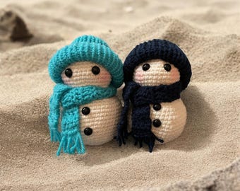May include: Two crocheted snowmen with blue and teal hats and scarves. The snowmen are sitting on a sandy surface.