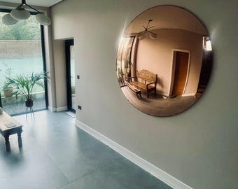 May include: A large, circular, bronze-tinted wall mirror mounted on a light gray wall. The mirror features a convex surface that reflects the interior space, including a wooden bench, a doorway, and a ceiling light fixture, creating a wide-angle view.