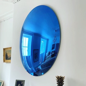 Puede incluir: Un gran espejo de pared de forma ovalada con una superficie reflectante de color azul vibrante. El espejo est&aacute; montado en una pared blanca lisa, creando un reflejo azul profundo y distorsionado del interior, incluyendo ventanas y l&aacute;mparas.