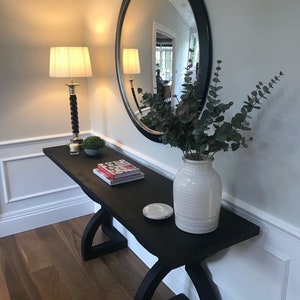 May include: A dark wood console table with a curved base, holding a white vase with greenery, a round mirror, a lamp, and decorative items. The table is made of wood and has a dark finish. The mirror has a black frame.