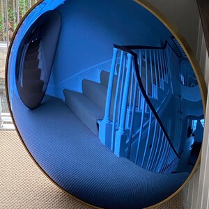 Puede incluir: Un gran espejo convexo circular con un marco de metal fino en tono dorado. La superficie reflectante est&aacute; tintada de azul y muestra un reflejo distorsionado y gran angular de una escalera interior con barandillas blancas.