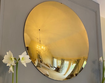 May include: A large, round, gold-colored mirror hangs on a gray wall. The mirror is convex, reflecting the room behind it. Two white amaryllis flowers in silver vases are on a white mantel below the mirror.
