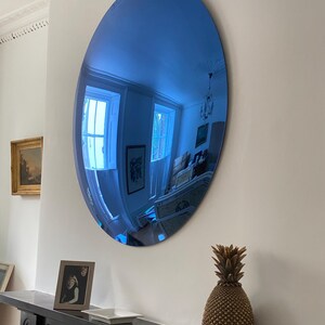 Puede incluir: Un gran espejo ovalado sin marco con una superficie reflectante de tono azulado. El espejo est&aacute; montado en una pared lisa y refleja la habitaci&oacute;n circundante, incluyendo ventanas, una l&aacute;mpara de techo y otros detalles arquitect&oacute;nicos.
