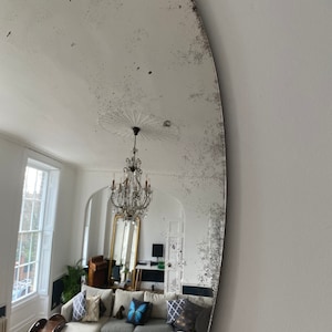 May include: A large oval mirror with a distressed silver finish reflects a living room with a white sofa, a chandelier, and a large window. The mirror has a unique, aged look.