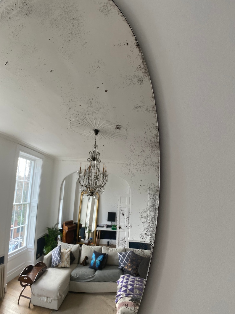 May include: A large oval mirror with a distressed silver finish reflects a living room with a white sofa, a chandelier, and a large window. The mirror has a unique, aged look.