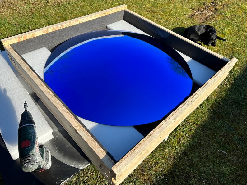 Puede incluir: Una gran superficie circular de color azul profundo y reflectante, encerrada en una caja de madera cuadrada. La caja est&aacute; forrada con espuma blanca para asegurar el objeto. Un taladro inal&aacute;mbrico est&aacute; colocado cerca de la esquina de la caja.