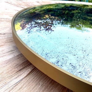 Pu&ograve; includere: Uno specchio rotondo con cornice dorata e una finitura leggermente usurata, in stile antico. Lo specchio riflette un'immagine sfocata di fogliame verde.