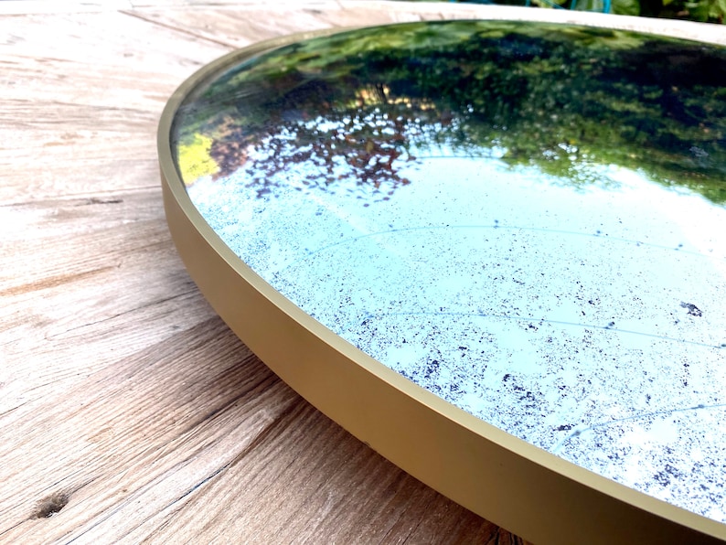 Pu&ograve; includere: Uno specchio rotondo con cornice dorata e una finitura leggermente usurata, in stile antico. Lo specchio riflette un'immagine sfocata di fogliame verde.