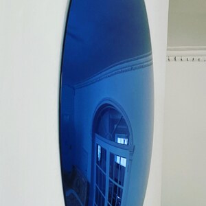 Puede incluir: Un espejo de pared grande de forma ovalada con una superficie reflectante te&ntilde;ida de azul intenso. El espejo est&aacute; montado verticalmente en una pared blanca lisa, mostrando un reflejo distorsionado de una puerta arqueada.