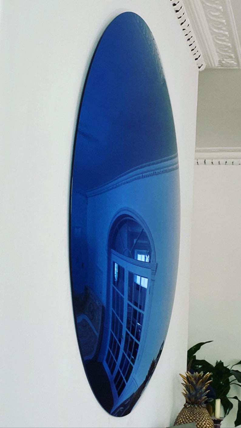 Puede incluir: Un espejo de pared grande de forma ovalada con una superficie reflectante te&ntilde;ida de azul intenso. El espejo est&aacute; montado verticalmente en una pared blanca lisa, mostrando un reflejo distorsionado de una puerta arqueada.