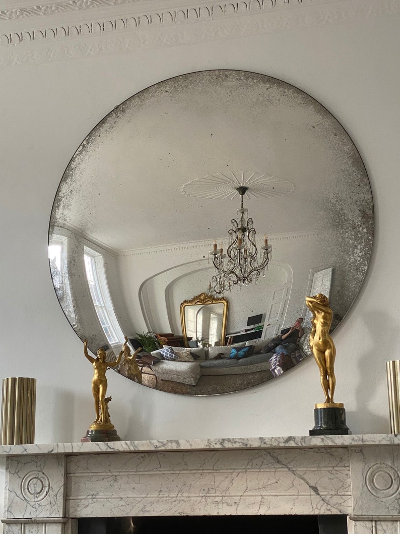 May include: A large, round, antique mirror with a distressed silver finish hangs above a marble fireplace mantel. Two gold-toned statues of figures stand on the mantel.
