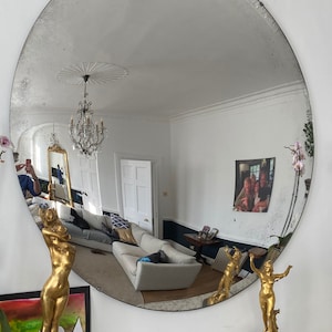 May include: A large, oval, antique mirror with a silver frame and a distressed finish. The mirror is reflecting a room with a white ceiling, a chandelier, and a white couch. There are two gold statues on the mantle below the mirror.