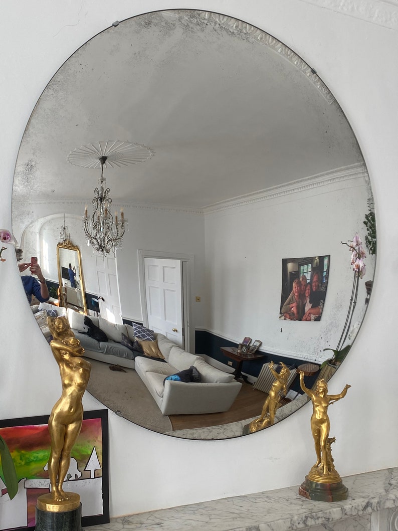 May include: A large, oval, antique mirror with a silver frame and a distressed finish. The mirror is reflecting a room with a white ceiling, a chandelier, and a white couch. There are two gold statues on the mantle below the mirror.